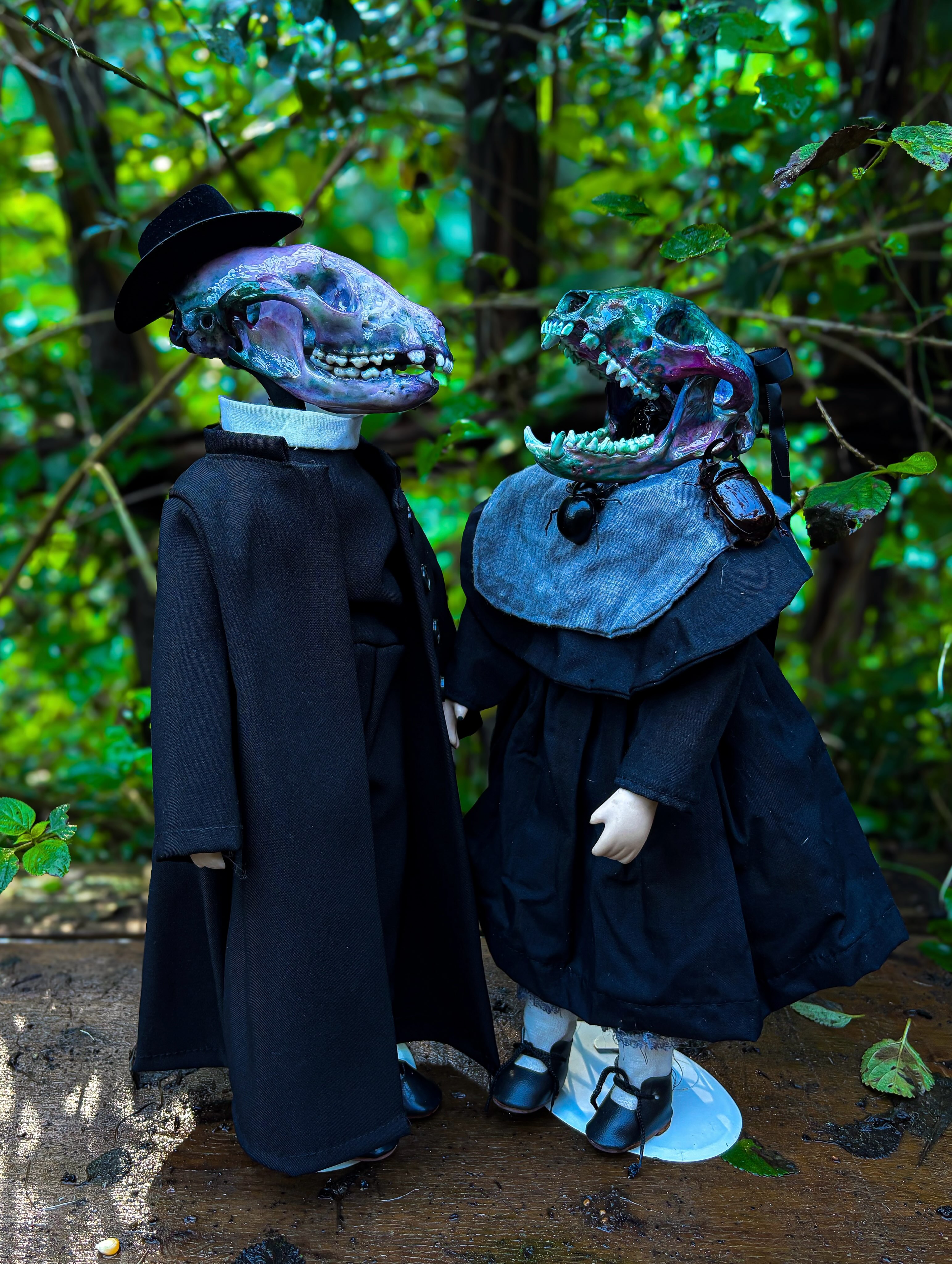Lucien and Bella — A pair of antique porcelain dolls with dyed raccoon skulls, dressed in dark, delicate finery.